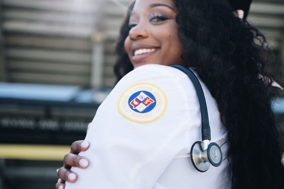 From below side view of crop smiling ethnic female in medical uniform showing emblem of Southern University School of Nursing on sleeve with dangling stethoscope over shoulder while standing on street in sunlight